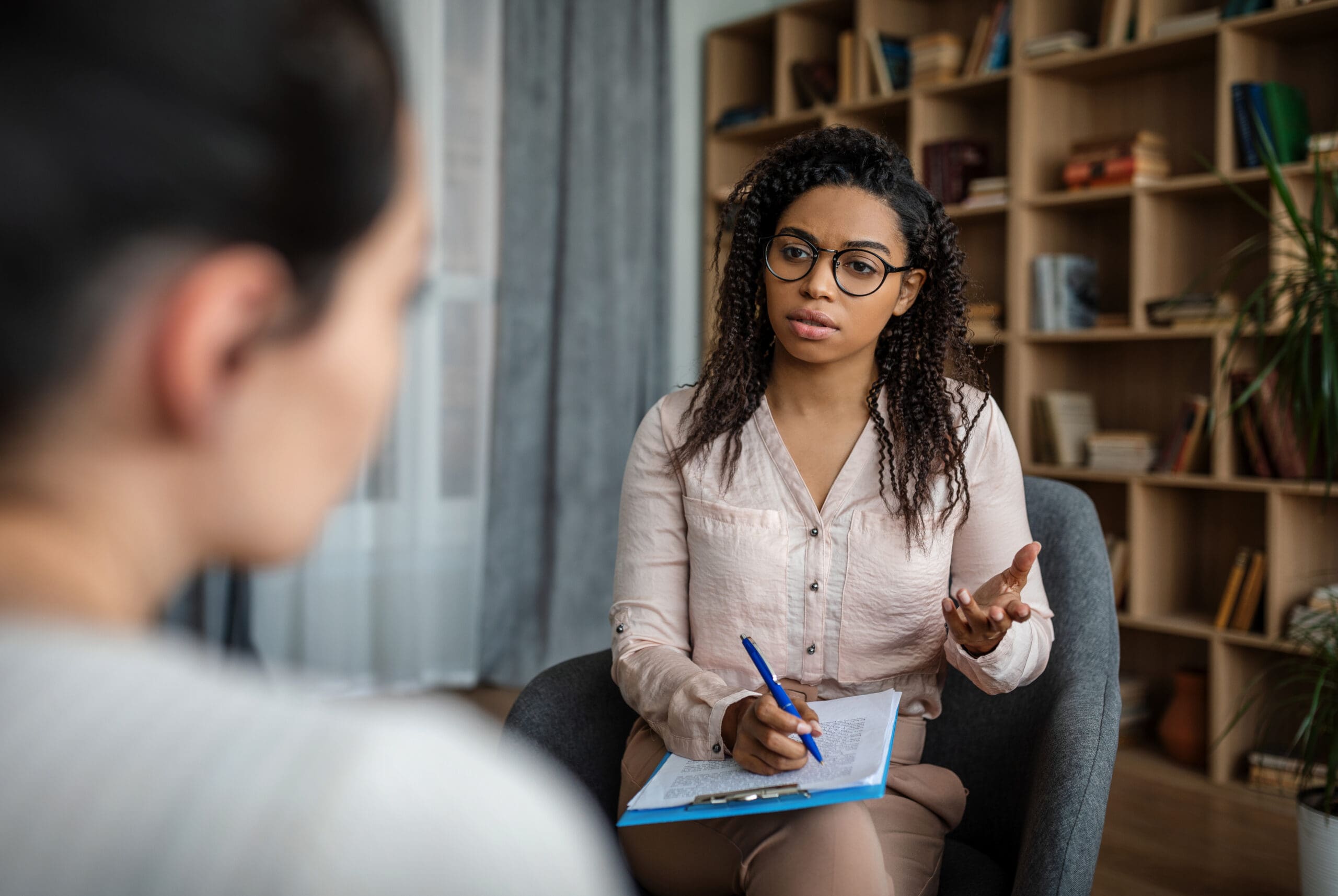 Serious young african american woman psychologist in glasses talking with european patient in office