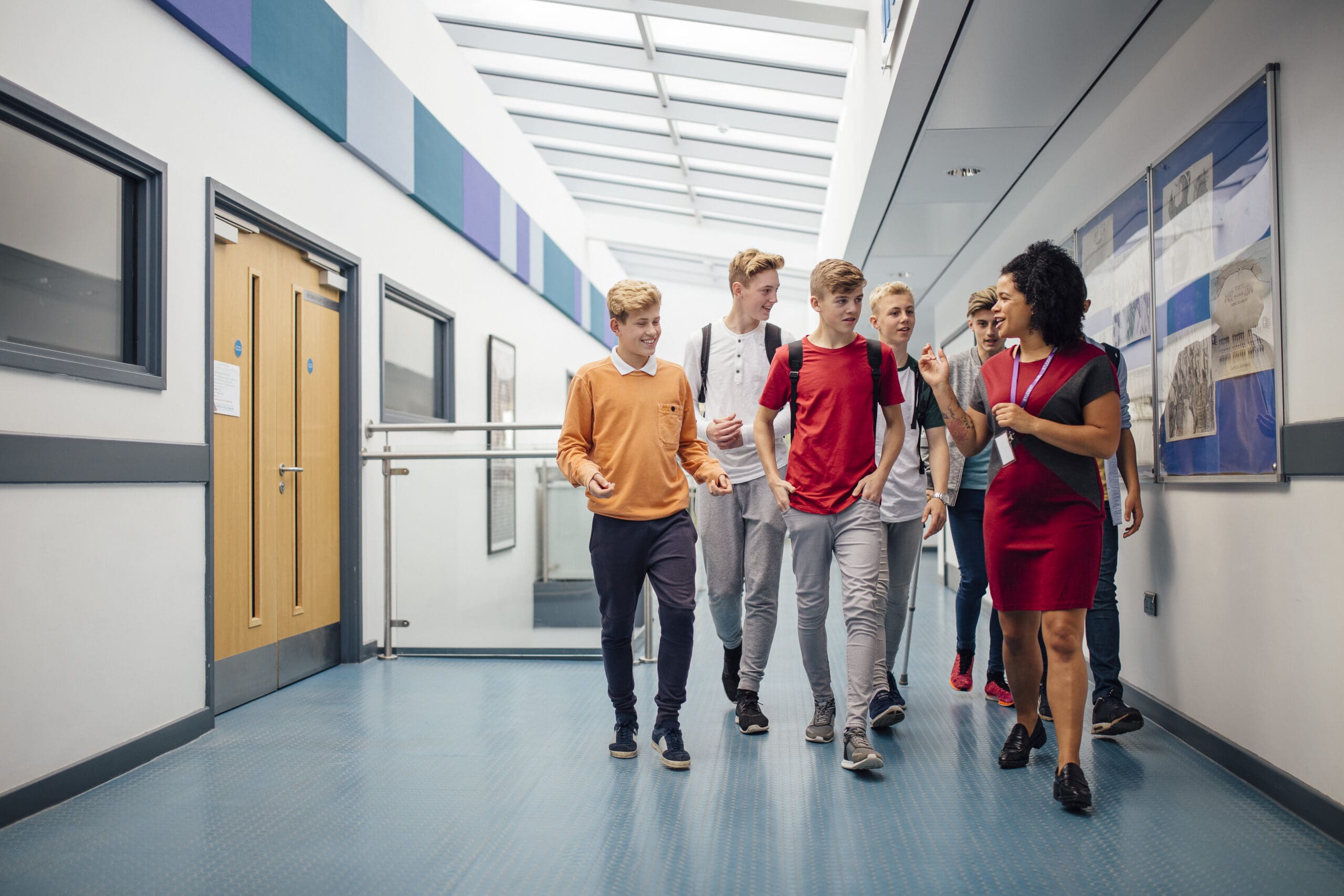 Female teacher is walking down the school hall with a group of male teenagers. She is giving them a tour of the school as it is their first day.