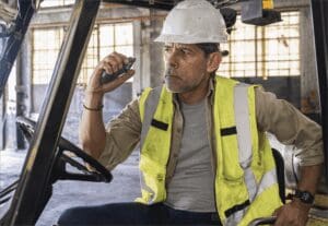 Focused worker in a hard hat and safety vest communicates using a Relay device while seated in a forklift inside an industrial facility.