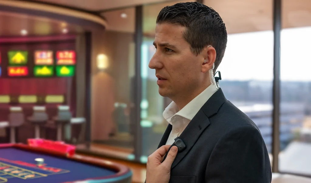 A casino security team member wearing an earpiece communicates using a Relay device near a gaming table and slot machines.