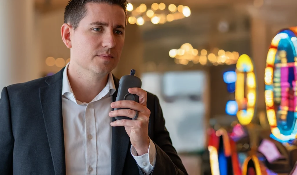 A casino security team member in a suit holds a Relay device while standing near brightly lit slot machines, appearing focused during communication.