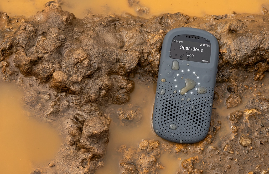 A Relay device labeled “Operations – Jon” lies in muddy water, showing its durability and clear screen despite being wet and dirty.