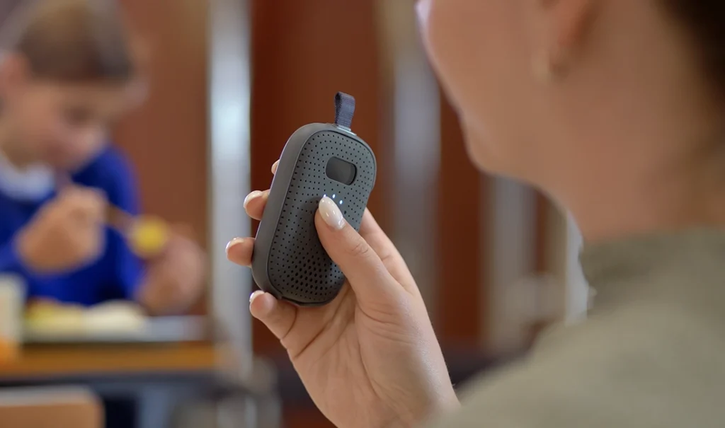 A school staff member holds a Relay device in a cafeteria, with a student eating in the background.