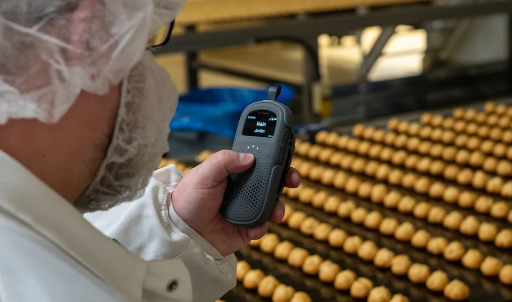 Food production worker uses RelayX device to communicate on the manufacturing floor near a conveyor of baked goods.