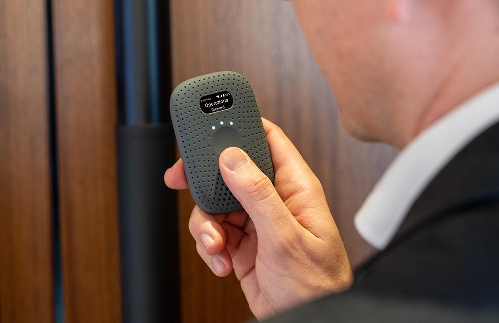 Person in a suit holding a gray RelayM device, pressing the button to send a message, with the screen displaying āOperations ā Richard.ā