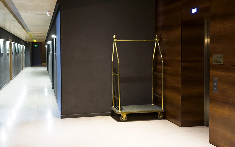 Empty brass hotel luggage cart positioned near an elevator with a “Staff Only” sign in a modern hotel hallway.