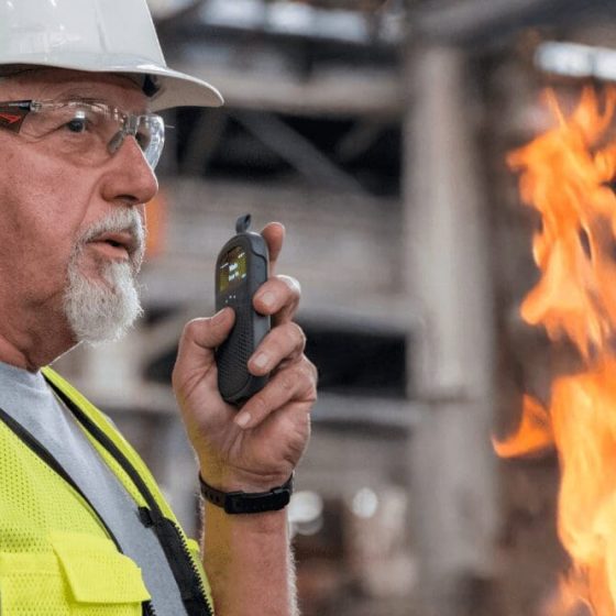 Older industrial worker wearing a hard hat and safety vest uses a Relay device to communicate near an active fire, highlighting emergency communication in hazardous environments.