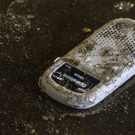 A Relay communication device covered in dirt and water lies on a wet, gritty industrial floor, displaying a message from “Operations – Vince” on the screen.