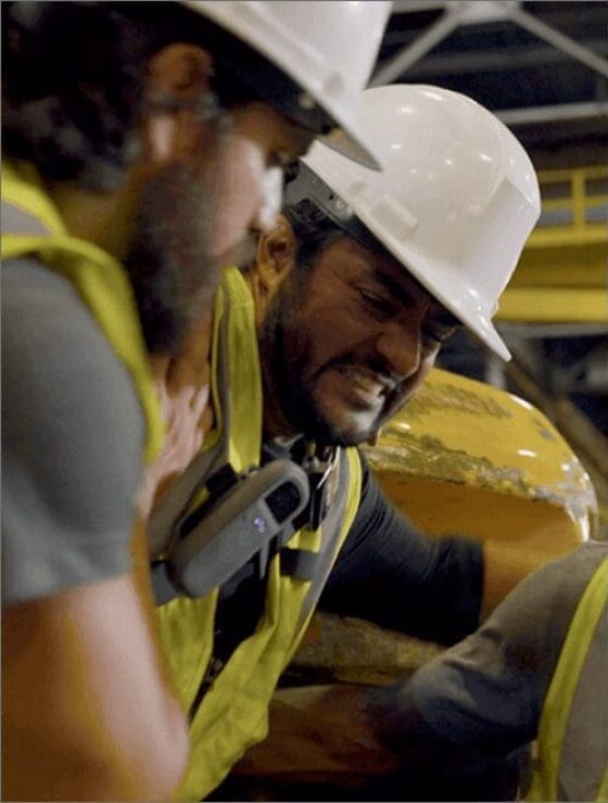 Industrial workers in safety vests and hard hats help and injured worker, with a Relay device visibly clipped to one worker’s vest.