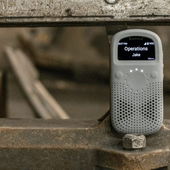 A Relay communication device rests on a piece of industrial equipment in a workshop setting, displaying a message from “Operations – Jake” on its screen.